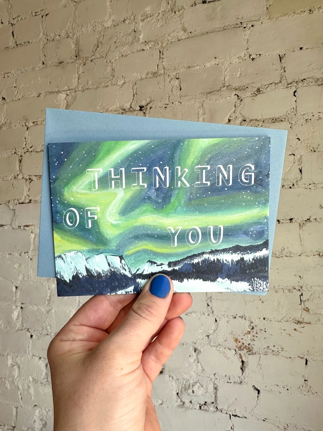 a greetings card featuring an illustration of the northern lights landscape with the text “thinking of you” with a blue envelope