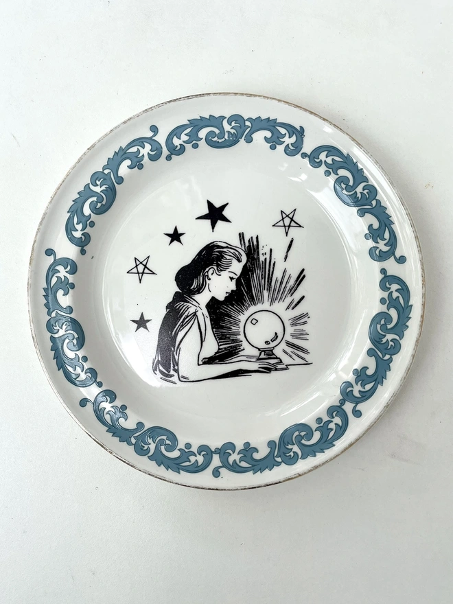a vintage plate with ornate border has a vintage illustration of a fortune teller and crystal ball in the centre