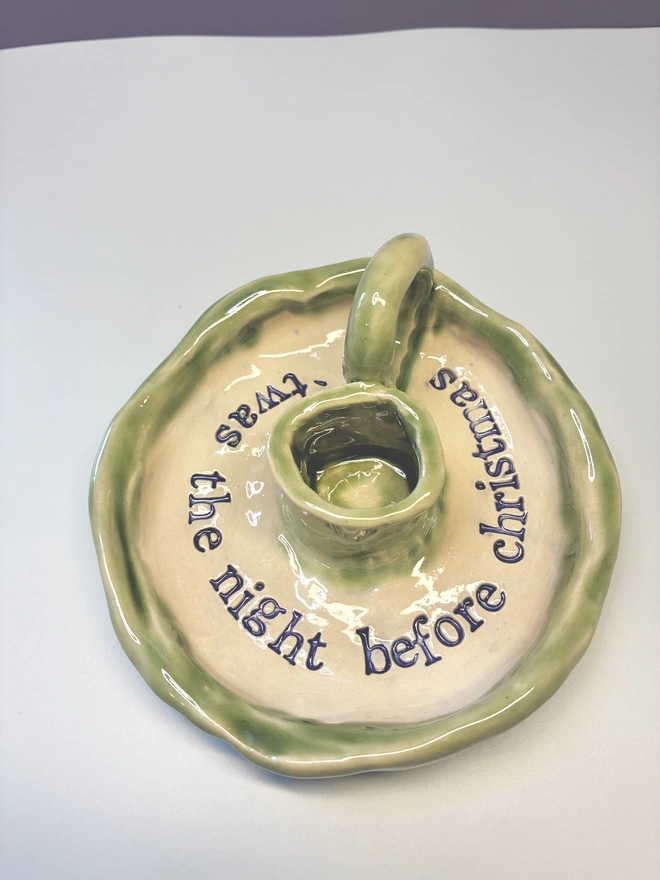 Handmade ceramic Christmas taper candlestick holder with a soft green glazed edge and the words “‘Twas the night before Christmas” in dark blue lettering, arranged around the candle holder opening. Photographed against a pale background.
