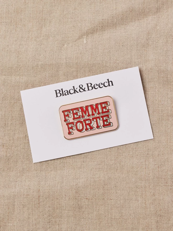 A calamine pink rectangular enamel pin with the words Femme Forte written in red 