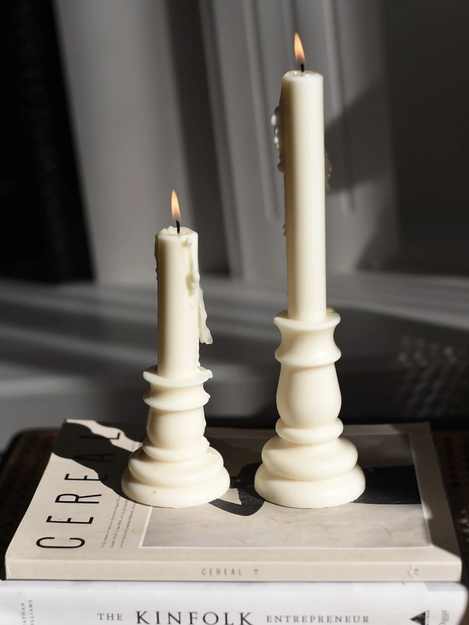 Baluster Christmas Candles (Set Of 2). Shadows of two candlestick candles with melted wax drips falling across a white panelled wall in low sunlight. The striking silhouettes show off the classic turned shape of these sculptural candles.