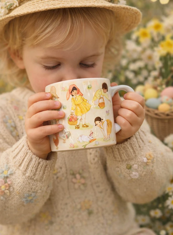 A photograph showing a small child drinking from an illustrated Easter mug. the mug features a vintage style wrap design showing little easter images a girl with a bunny bonnet, a girl with a bunny and a carrot and a little boy easter egg hunting with a basket