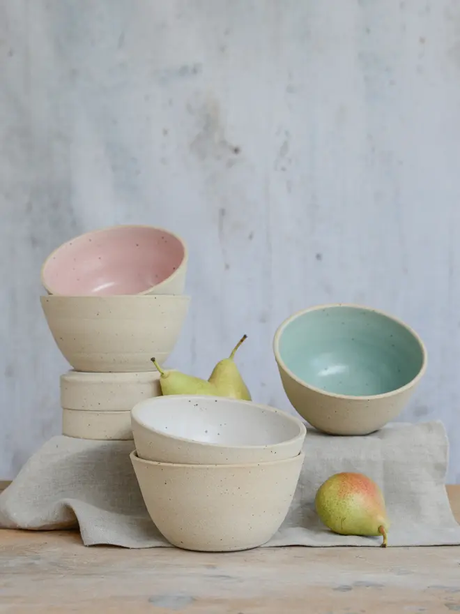 Stacks of pink, white and a mint green cereal soup at different levels on top of a linen cloth on a wooden surface. There are decorative pears arounds the bowls.