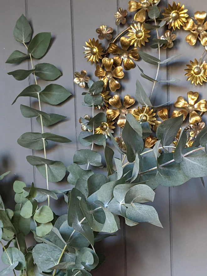 eucalyptus in vase with metallic floral wreath seen in the background on grey wall