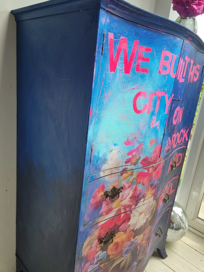 A vintage tallboy cabinet painted in vibrant blues, neons and flowers with the text that reads 'we built this city on rock and roll' in neon pink.