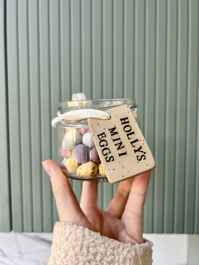 A Jar filled with mini chocolate eggs has a brown speckled ceramic tag with the wording 'Holly's Mini Eggs' in black lettering