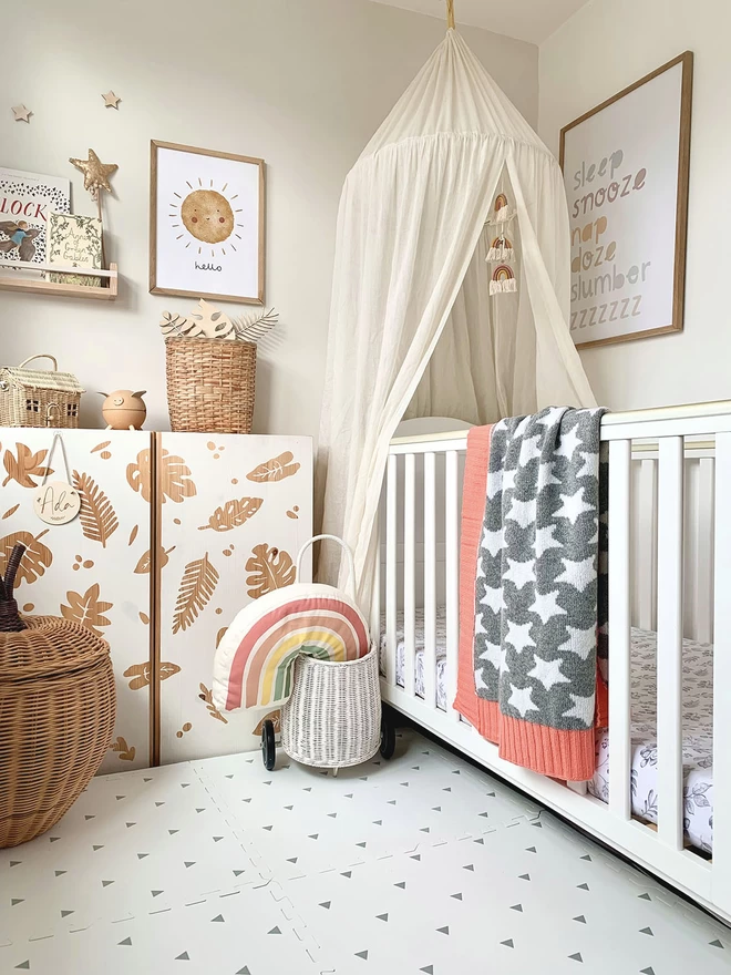 A neutral scandi style nursery with lots of natural wood, wicker accessories and a white cot. Over the cot is draped a grey and white star blanket with coral pink trim. This shot shows the reverse of the blanket with grey background and white stars. A poster on the wall features typography words describing sleep in muted rainbow colours.
