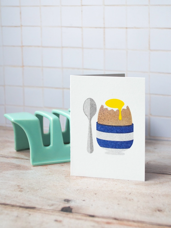 The breakfast table is all set with toast rack and a mini risograph printed greeting card featuring a delightful illustration of a soft boiled egg in a blue and white striped egg cup with a small spoon.