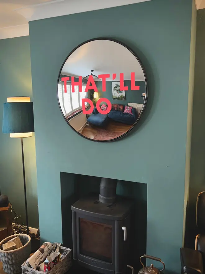 Round convex mirror with 'That'll Do' handpainted in neon pink, on a teal fireplace wall above a log burner, with a tall lamp to the side