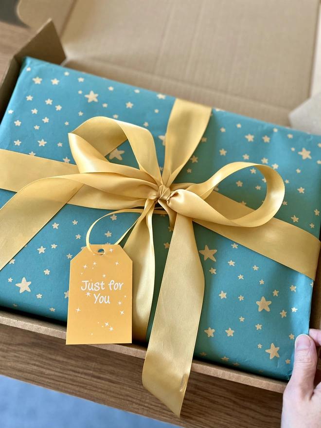 Rectangular box gift wrapped in blue paper with yellow stars and tied with a large gold bow & yellow gift card saying 'Just for You', lying in a neat fitting kraft postal box. 