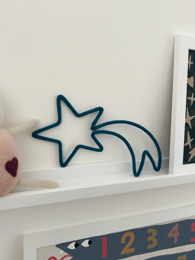 A knitted wire sign in the shape of a shooting star propped on a picture shelf in a kid's room.