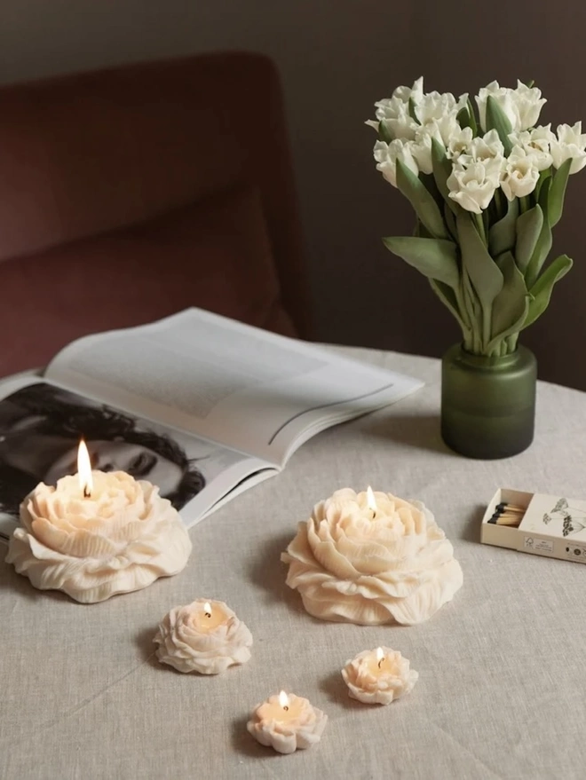 Large White Peony Candle. Lit peony candles in large and small sizes glowing on a linen tablecloth, with an open magazine, white tulips in a green vase and a box of matches. The set of floral candles creates a soft, cosy atmosphere for relaxing at home.