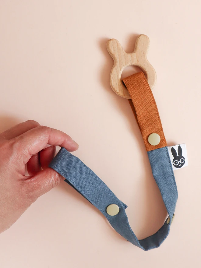 Handmade toy straps in woodland shades, featuring sturdy snap fastenings and small fabric tags with a bunny logo, attached to a bunny shaped wooden teether on on side and held by a hand on the other side.