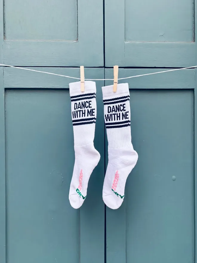 Socks with DANCE WITH ME text shown hung by clothes pegs on washing line in front of greyish blue shutters