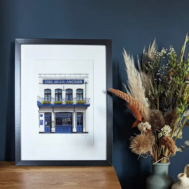 Hammersmith The Blue Anchor Giclée Print (Unframed), a print of a blue pub sitting in a black frame on a wooden surface against a navy blue backdrop.  