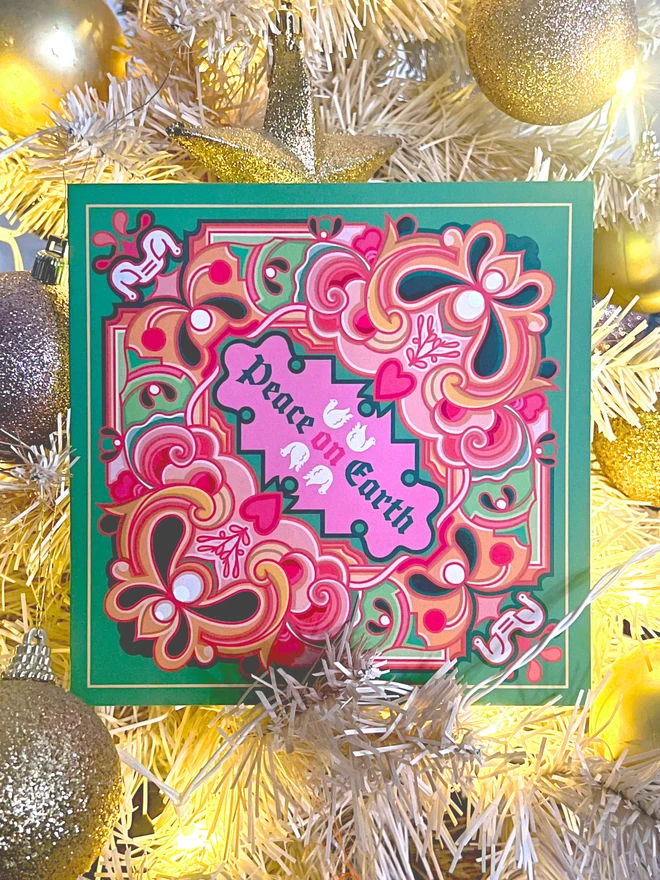 An abstract Christmas card design with Peace on Earth at the centre with drawings of two doves above and two below, surrounded by a multi-coloured design on a green background. The card features greens, pinks and oranges and rests in white and gold tinsel and baubles.