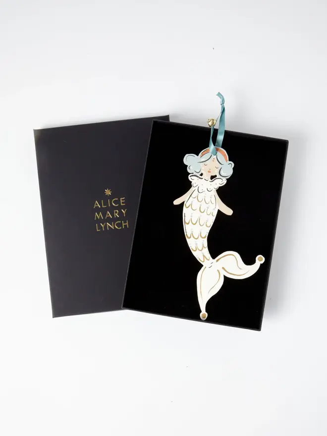 Mermaid Tree Dancer. the mermaid tree dancer hanging ornament in cream and gold tones sits inside its black presentation box, with the lid open to reveal "alice mary lynch" embossed in gold lettering. the mermaid figure has pale blue curled hair and an orange headband, with a cream scalloped scale-patterned tail edged in gold, curling into a jester-style pointed tip. the branded box gives the piece a polished, gift-ready presentation.