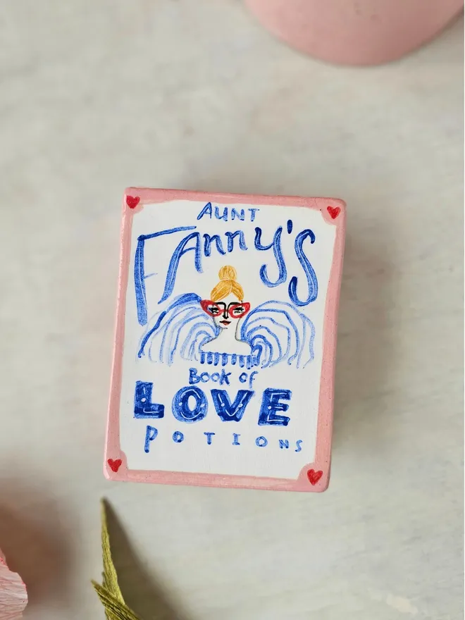  A charming ceramic trinket box designed to look like a vintage book, painted in shades of pink and red. The cover features the title "Aunt Fanny’s Book of Love Potions" in a playful, illustrated style. With its book-like shape and whimsical design, this decorative piece adds a touch of romance and mystery while offering a secret spot to store small treasures.