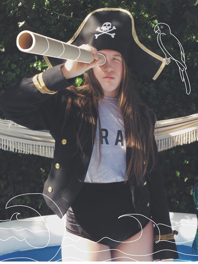 Model wearing a pirate hat with gold trim and white skull and cross-bone print, a pirate eye patch and grey PIRATE T-shirt. Looking through a telescope with a hand drawn parrot on her shoulder