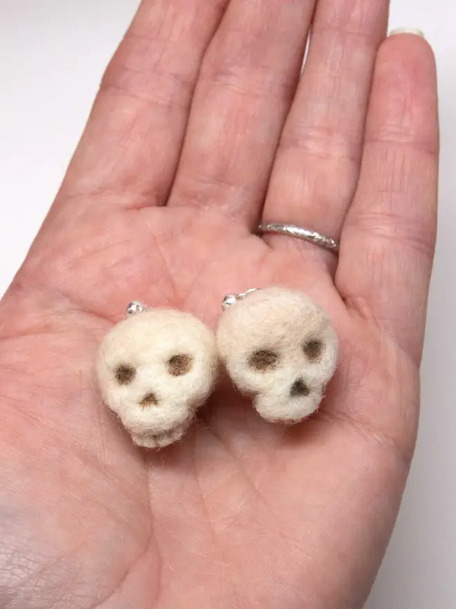 Two needle-felted skull earrings in the palm of a hand. Made by the artist Laura Cronin aka Bumble & Earwig