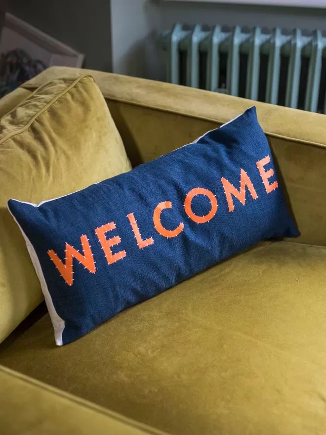 Welcome cushion by Love Welcomes seen on a yellow sofa.
