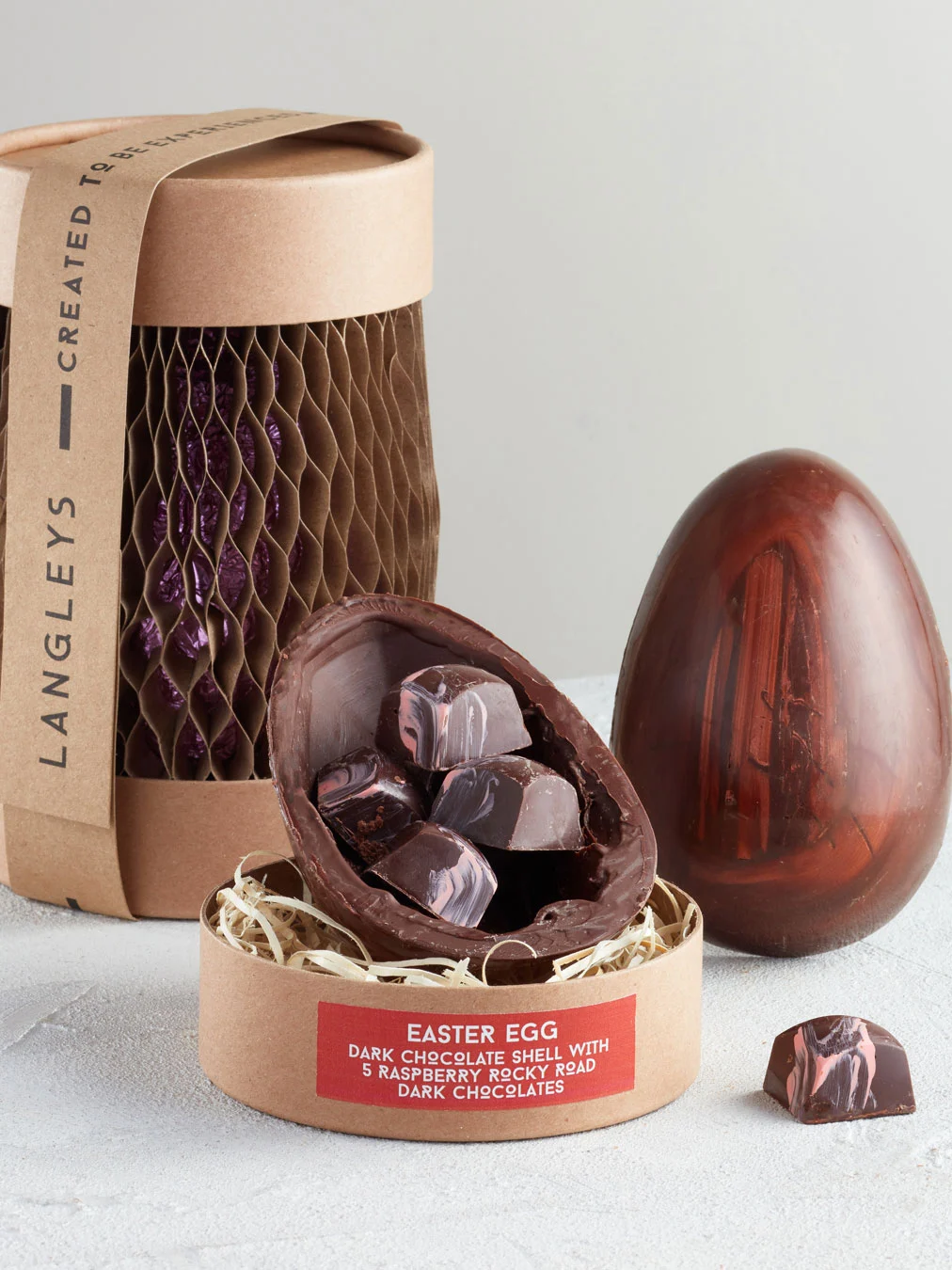 Dark Chocolate Easter Egg With Raspberry Rocky Road, a chocolate egg sitting on a white surface. It is accompanied by another chocolate egg that is opened showing the chocolates within. 