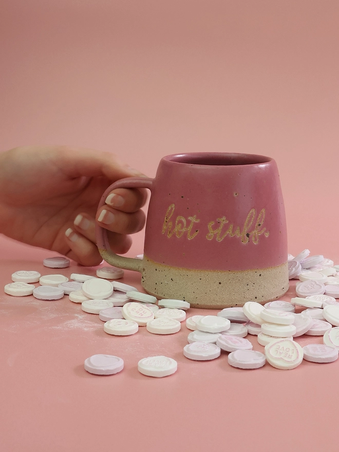 text is written on the front of a pink handmade mug which reads hot stuff, the mug sits infront of a paler pink background 