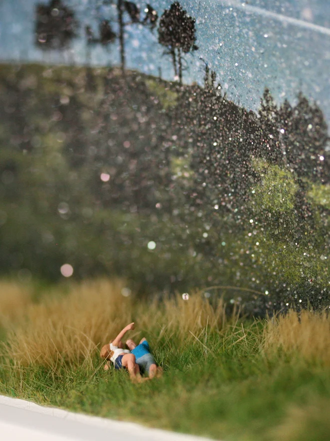 Miniature scene in an artbox showing a tiny man and woman lying in the grass watching a shooting star, against a dramatic star-filled glittery night sky.  