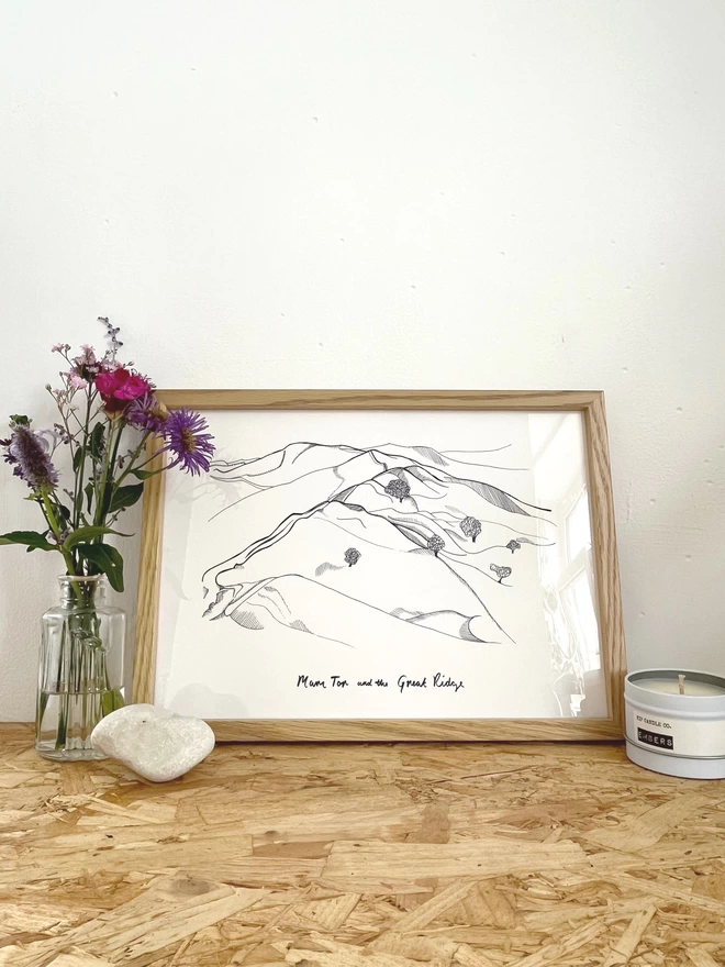 a print featuring an fine line illustration of Mam Tor and the Ridge Walk in the peak district in a frame next to some flowers, a rock and a candle