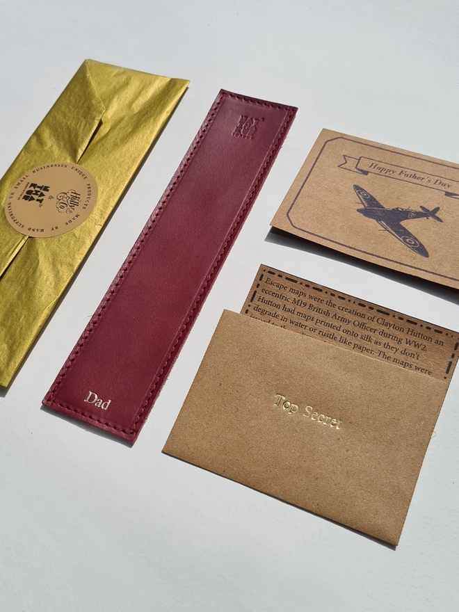 a wine red silk escape leather bookmark personalised with dad in gold foil for fathers day gift packaged in gold tissue paper with a free gift card with a spitfire airplane