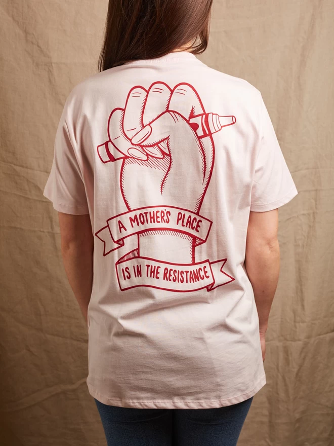 Model is showing the back of the Black & Beech pink A Mother’s Place is in the resistance t-shirt. The red print on the back shows a fist clutching a crayon and the with the words A Mother’s Place is in The Resistance written on a ribbon wrapped around the wrist 