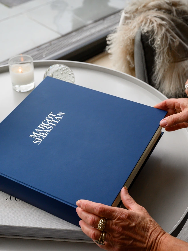 on a grey coffee table there's a large blue photo album personalised with two names on the front. 