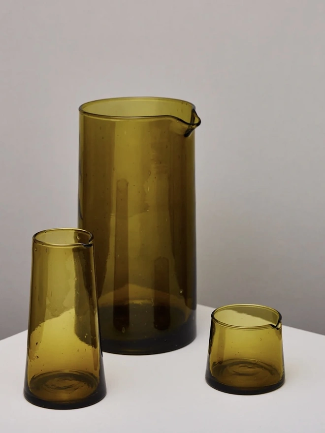 Glass Carafe, a large amber glass jug sitting on a plain counter top against a grey backdrop. It is accompanied by other amber drinking vessels