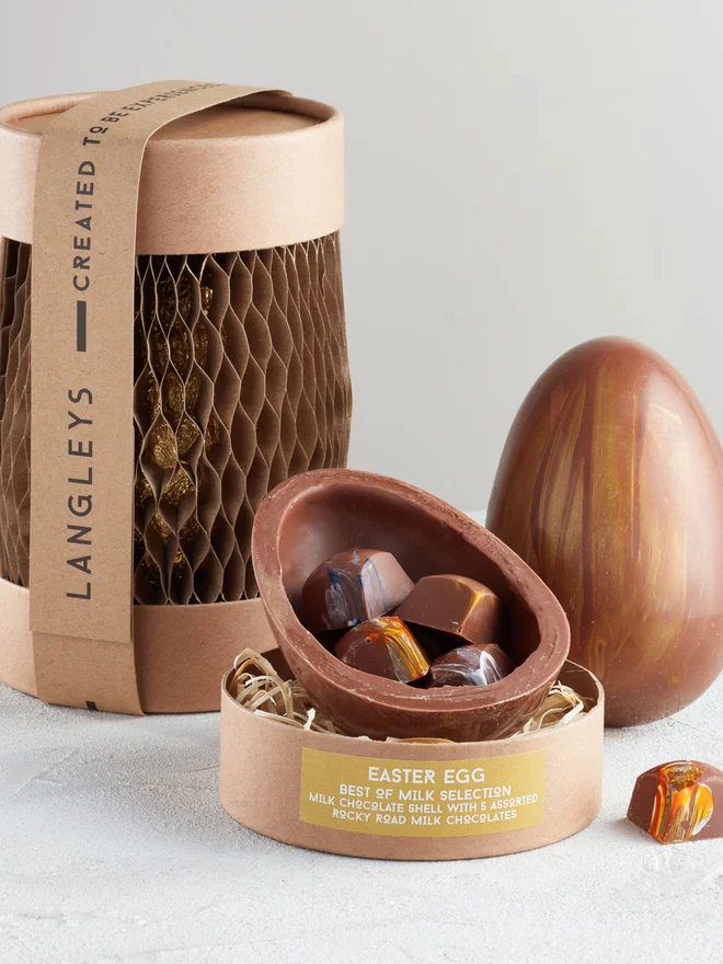 Best Of Milk Chocolate Easter Egg, a chocolate egg sitting on a white surface. It is accompanied by another chocolate egg that is opened showing the chocolates within. 