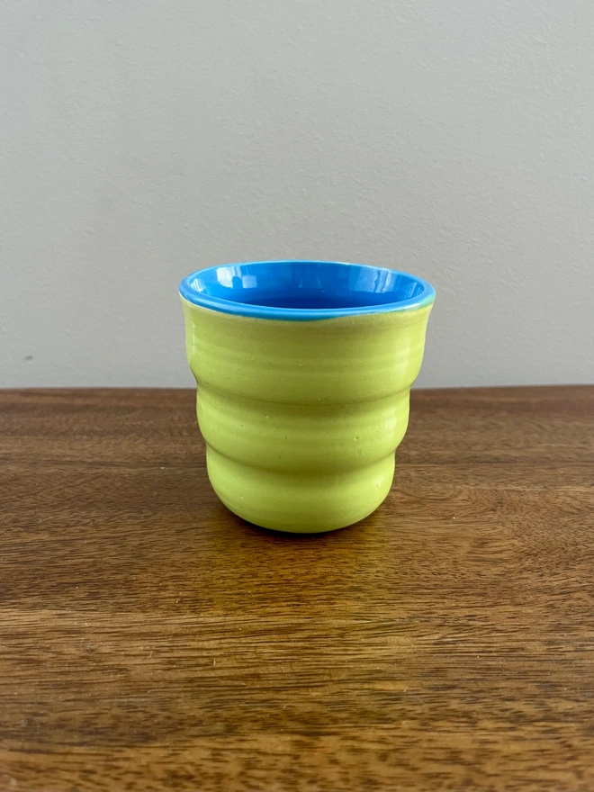 mini ceramic tumbler with wavy sides on a wooden table. the tumbler has a lime green glaze on the outside and azure blue on the inside and rim. 
