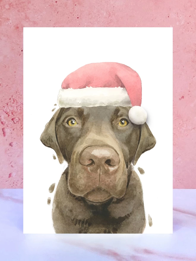 A Christmas card featuring a hand painted design of a chocolate labrador, stood upright on a marble surface surrounded by pompoms. 