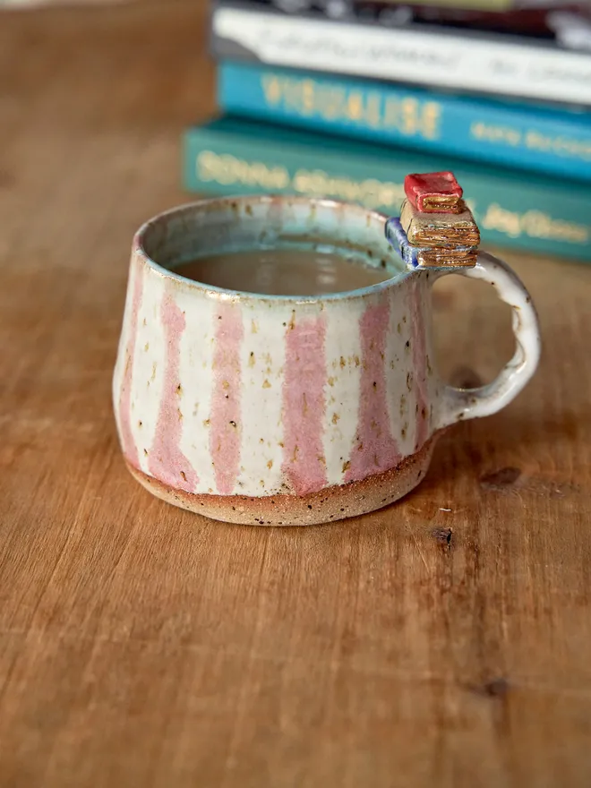 Pink and white striped ceramic mug with book detail on the handle, pictured on a wooden table with a pile of books in the background