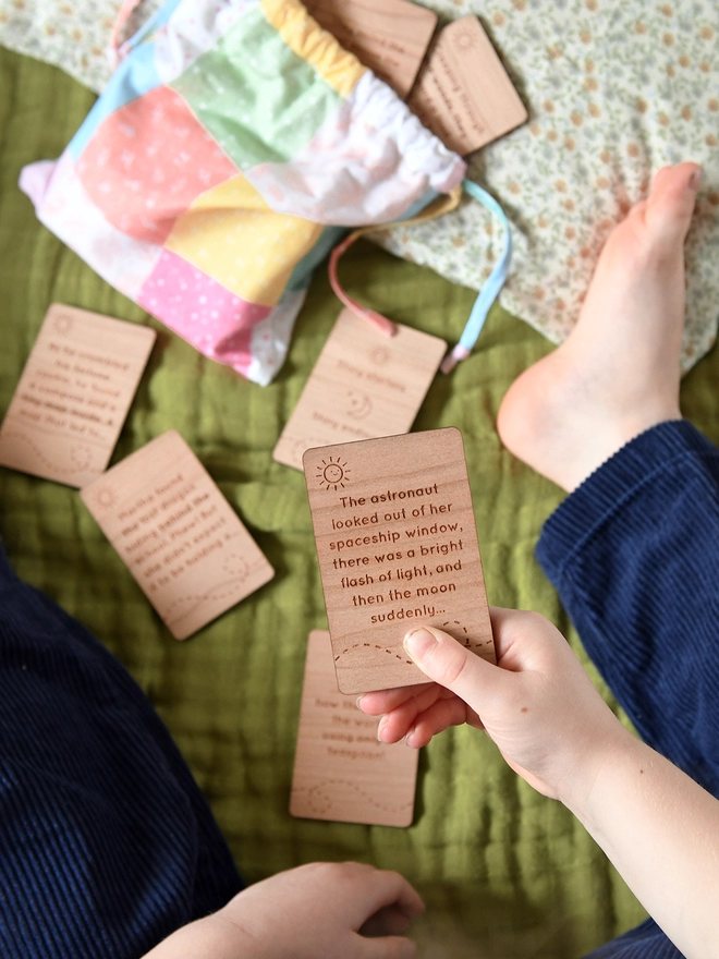 A young child holds a wooden story card with engraved wording in her hand above a green blanket. Several other cards lay on the blanket.