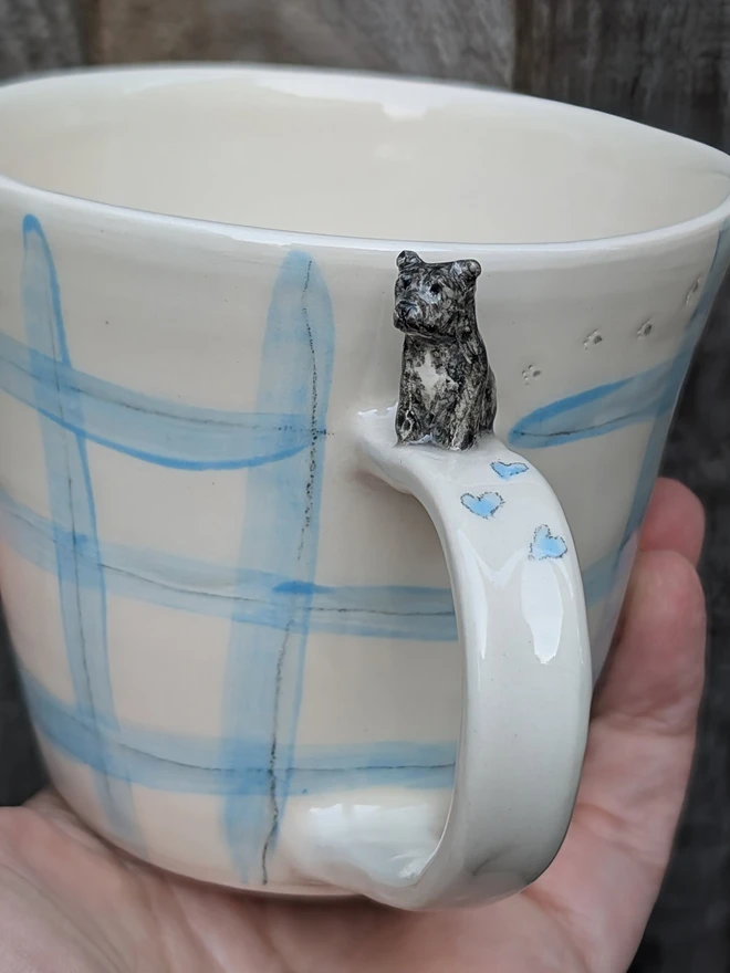 personalised dog mug with a tiny Staffordshire bull terrier on the handle, tiny paw prints and blue gingham check and hearts held in a hand