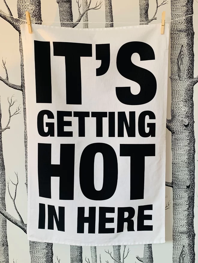London Drying Its Getting Hot in Here in black text on white tea towel hanging washing line style in front of woods pattern wallpaper