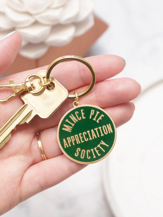 Hand holding out a Green enamel keyring featuring 'Mince Pie Appreciation Society' design