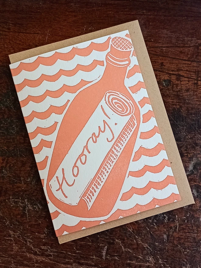 Card with envelope folded inside. Printed on front: Cartoon style, flat colour, light red. Wavy pattern with matte bottle outline containing scroll with text, "Hooray!"