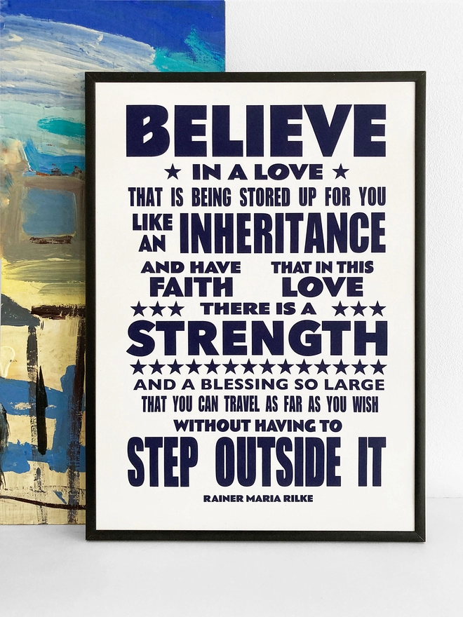 Framed typographic print with words by Rainer Maria Rilke. The quote reads “Believe in love that is stored up for you like an inheritance and have faith that in this love there is a strength and a blessing so large that you can travel as far as you wish without having to step outside it” The print rests against a blue and yellow abstract painting.