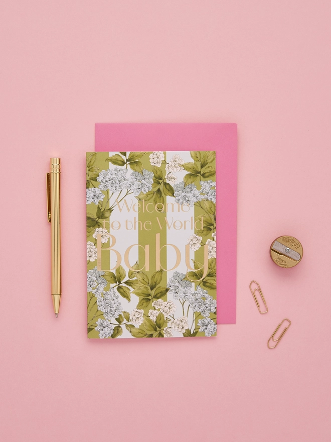 'Welcome to the World baby' card with gold foil wording and illustrated blue hydrangea flowers on a green and light blue striped base with a hot pink envelope