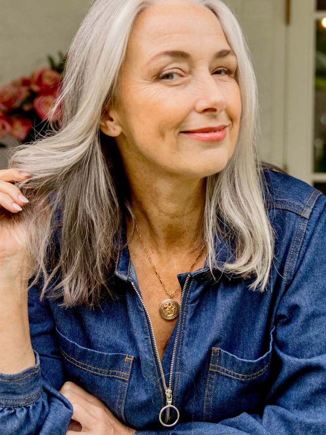 Woman with grey hair in a denim jumpsuit smiling and wearing a gold paperclip chain with a round lucky 13 charm