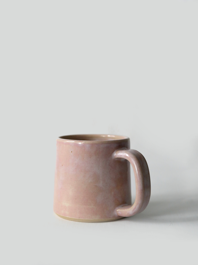 A handmade ceramic standard mug in glossy blush pink glaze with a chunky loop handle, photographed against a plain light grey background
