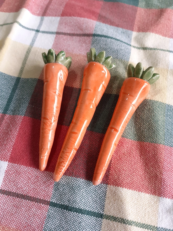 Close-up of three handmade ceramic carrot candleholders laid on a pastel check tablecloth, showing glossy glaze and sculpted leafy tops.