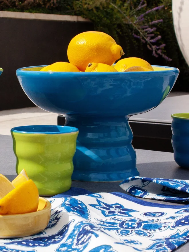 a colourful pedestal bowl filled with lemons. the bowl has a wide shallow bowl and a chubby elevated foot. The bowl is bright azure blue on the outside and lime green on the inside. The bowl is sitting on a patio table surrounded by a tablecloth and matching tumblers