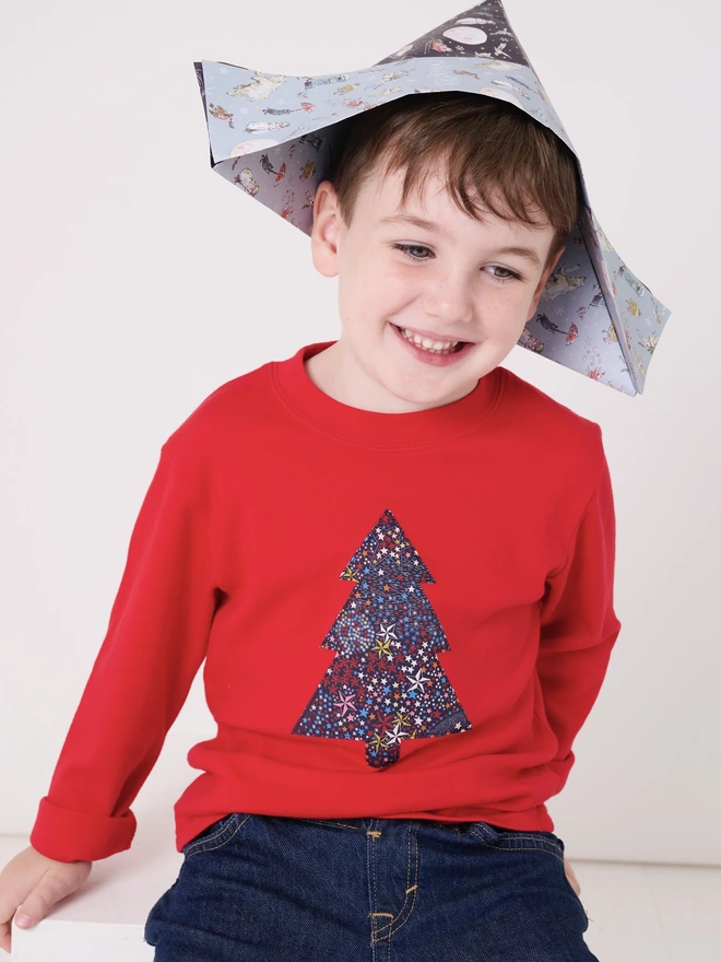 A smiling boy wearing a red t-shirt with a starry print Christmas tree sewn on the front
