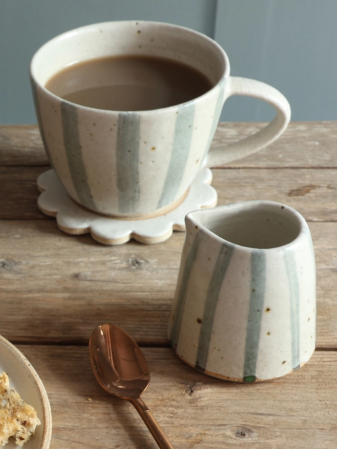 Handmade Ceramic Stoneware Small Jug in Blue and White Stripe sat on a wooden surface in front of a mug in the same style filled with coffee. The mug sits on top of a handmade flower coaster. A brass spoon and a plate of cookies can be seen in the foreground. 